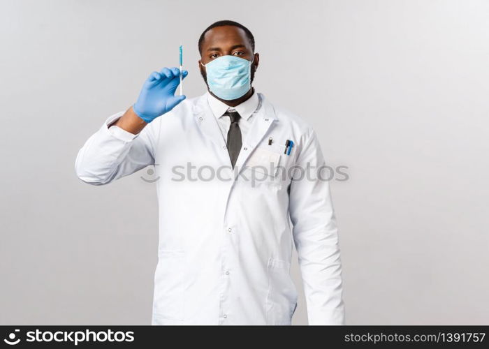 Covid-19, coronavirus patient treatment and laboratory concept. Serious professional african-american doctor ready to cure, holding syringe with blue vaccine, wear face mask and latex gloves.. Covid-19, coronavirus patient treatment and laboratory concept. Serious professional african-american doctor ready to cure, holding syringe with blue vaccine, wear face mask and latex gloves