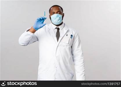 Covid-19, coronavirus patient treatment and laboratory concept. Serious professional african-american doctor ready to cure, holding syringe with blue vaccine, wear face mask and latex gloves.. Covid-19, coronavirus patient treatment and laboratory concept. Serious professional african-american doctor ready to cure, holding syringe with blue vaccine, wear face mask and latex gloves