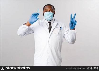 Covid-19, coronavirus patient treatment and laboratory concept. Happy african-american doctor treating people in hospital, show peace sign and hold syringe with blue vaccine, wear face mask.. Covid-19, coronavirus patient treatment and laboratory concept. Happy african-american doctor treating people in hospital, show peace sign and hold syringe with blue vaccine, wear face mask