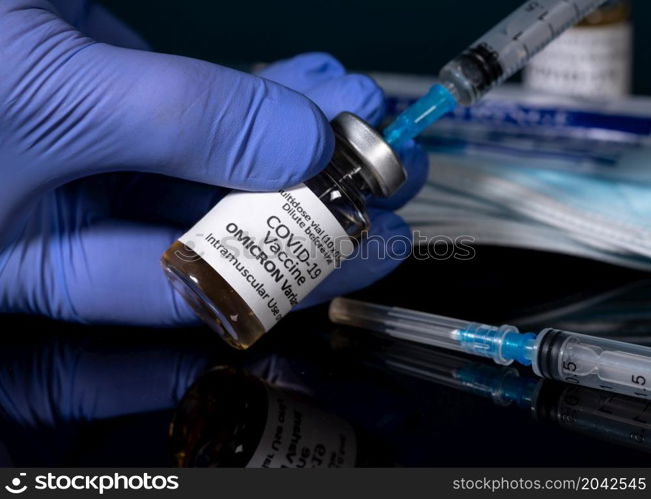 Covid-19 coronavirus vaccine developed for Omicron variant held by gloved hand with hypodermic syringe needle and new syringes in background. Close up of bottle of new Covid-19 vaccine for Omicron variant wtih gloved hand