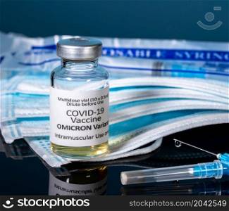 Covid-19 coronavirus vaccine developed for Omicron variant with hypodermic syringe needle with masks and new syringes in background. Close up of bottle of new Covid-19 vaccine for Omicron variant
