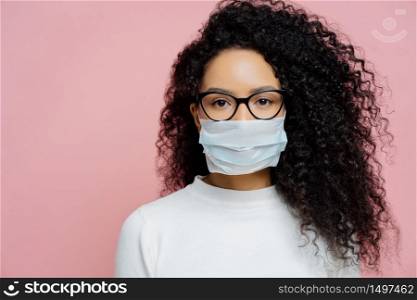 Covid-19, infectious virus. Close up shot of young woman with curly bushy hair, wears transparent glasses and medical disposable mask, cares about her health, protects in dangerious situation