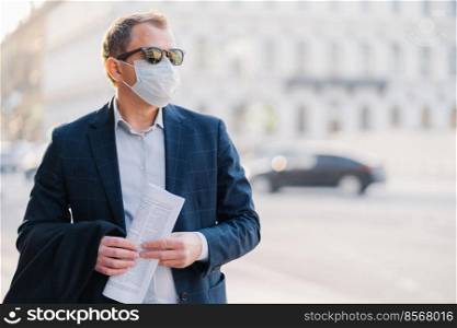 Covid-19. Man worker dressed in formal wear, holds newspaper in hands, poses at street, wears surgical mask, avoids crowded street not to catch virus, poses against blurred background with transport