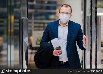 Covid-19, pandemic coronavirus concept. Office worker leaves work, wears medical face mask for infectious disease spread, dressed formally, holds newspaper and smartphone. Health and safety concept