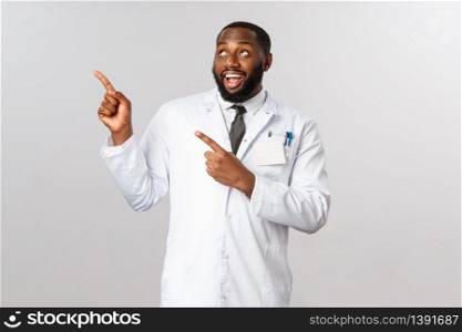Covid19, healthcare and clinic concept. Charismatic, handsome african-american male doctor, physician or pharmacy clerk pointing fingers upper left corner with pleased smile, recommend pills.. Covid19, healthcare and clinic concept. Charismatic, handsome african-american male doctor, physician or pharmacy clerk pointing fingers upper left corner with pleased smile, recommend pills