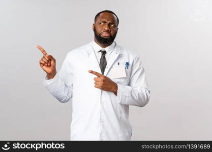 Covid19, healthcare and clinic concept. Portrait of confused and displeased, judgemental african-american doctor, grimacing in aversion, cringe looking and pointing left, patient not wearing mask.. Covid19, healthcare and clinic concept. Portrait of confused and displeased, judgemental african-american doctor, grimacing in aversion, cringe looking and pointing left, patient not wearing mask