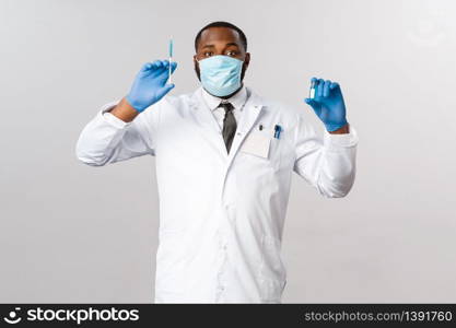 Covid19, pandemic and healthcare concept. Cheerful handsome african-american doctor showing vaccine in ampule and syringe, ready to tream patients, prepared for hospital check-up, grey background.. Covid19, pandemic and healthcare concept. Cheerful handsome african-american doctor showing vaccine in ampule and syringe, ready to tream patients, prepared for hospital check-up, grey background