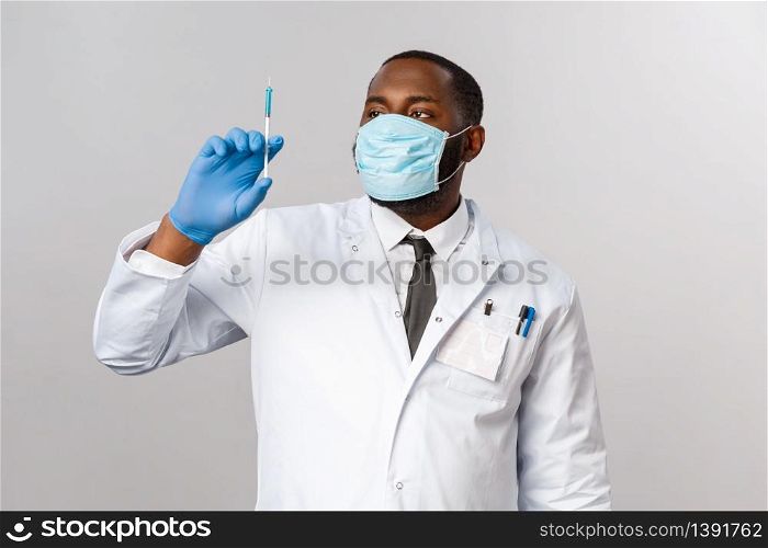 Covid19, pandemic and healthcare concept. Serious handsome african-american doctor in face mask, latex mask, looking at syringe with virus vaccine, treating patients, grey background.. Covid19, pandemic and healthcare concept. Serious handsome african-american doctor in face mask, latex mask, looking at syringe with virus vaccine, treating patients, grey background