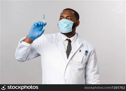 Covid19, pandemic and healthcare concept. Serious handsome african-american doctor in face mask, latex mask, looking at syringe with virus vaccine, treating patients, grey background.. Covid19, pandemic and healthcare concept. Serious handsome african-american doctor in face mask, latex mask, looking at syringe with virus vaccine, treating patients, grey background