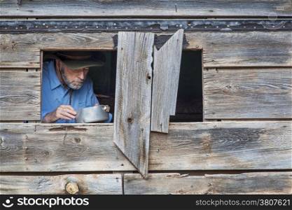 cowboy cooking or eating in an old shack, a face visible through window
