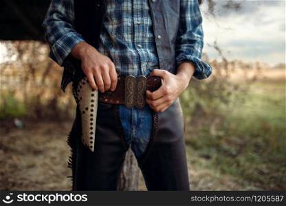 Cowboy in jeans and leather clothes with his hand on the revolver, texas ranch on background, western. Vintage male person with gun, wild west lifestyle