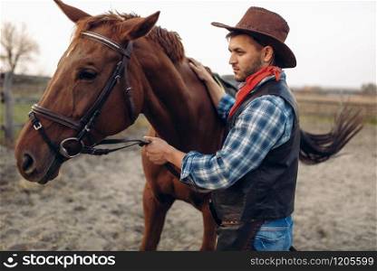 Cowboy in jeans and leather jacket poses with horse on texas ranch, western. Vintage male person with animal, wild west