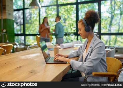 Coworking space concept. Young business woman freelancer wearing headphones using laptop computer for online work and video conferencing at shared open workplace. Young woman in headphones using laptop for online work