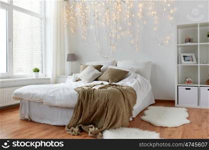 coziness, comfort, interior and holidays concept - cozy bedroom with bed and christmas garland lights at home