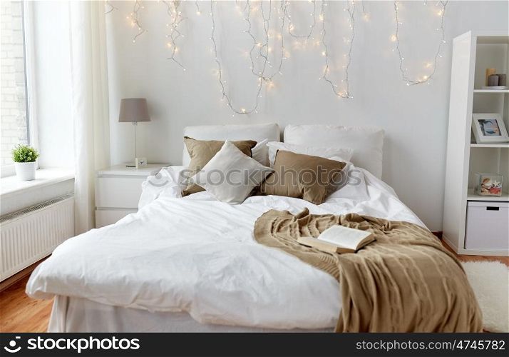 coziness, comfort, interior and holidays concept - cozy bedroom with bed and christmas garland lights at home
