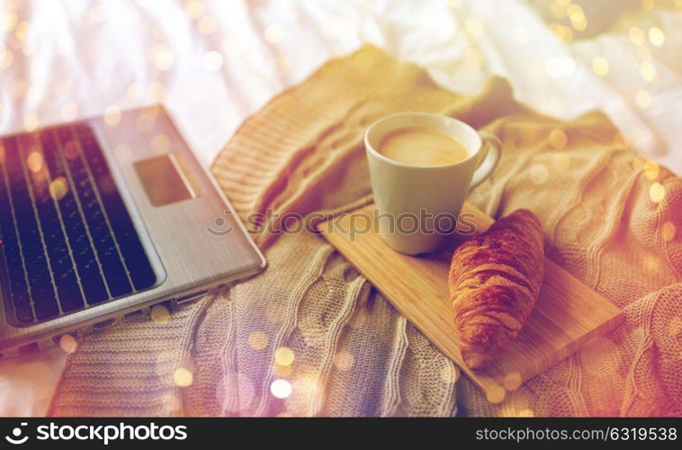 coziness, morning, technology and breakfast concept - bedroom with laptop computer, coffee cup and croissant on bed at home. laptop, coffee and croissant on bed at cozy home