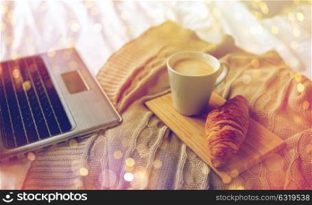 coziness, morning, technology and breakfast concept - bedroom with laptop computer, coffee cup and croissant on bed at home. laptop, coffee and croissant on bed at cozy home