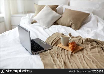 coziness, technology, interior and winter concept - cozy bedroom with laptop computer, coffee cup and croissant on bed at home