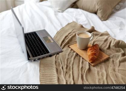 coziness, technology, interior and winter concept - cozy bedroom with laptop computer, coffee cup and croissant on bed at home