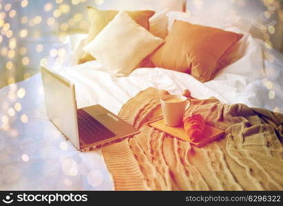 coziness, technology, interior and winter concept - cozy bedroom with laptop computer, coffee cup and croissant on bed at home. laptop, coffee and croissant on bed at cozy home