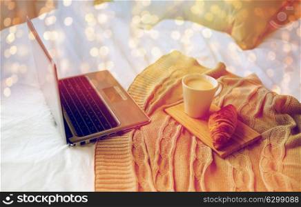 coziness, technology, interior and winter concept - cozy bedroom with laptop computer, coffee cup and croissant on bed at home. laptop, coffee and croissant on bed at cozy home