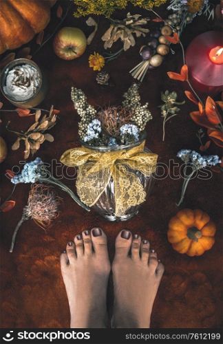 Cozy autumn mood at home. Female feet and vase with dried flowers arrangement on carpet floor with pumpkins, candles, and fall leaves. Top view