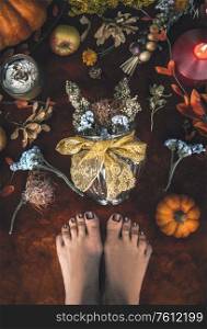 Cozy autumn mood at home. Female feet and vase with dried flowers arrangement on carpet floor with pumpkins, candles, and fall leaves. Top view