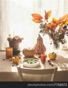 Cozy autumn still life. Sunny table setting with various pumpkins, autumn leaves bunch , candles, basket, plate and cutlery at window background with curtains and natural sunlight. Front view