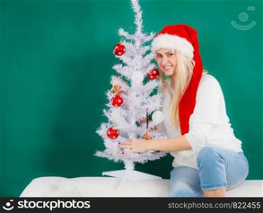 Cozy holiday interior, seasonal accessories concept. Blonde young woman in Santa hat decorating Christmas tree. Woman in Santa hat decorating Christmas tree