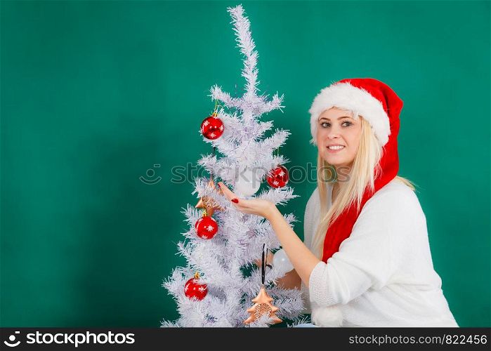 Cozy holiday interior, seasonal accessories concept. Blonde young woman in Santa hat decorating Christmas tree. Woman in Santa hat decorating Christmas tree