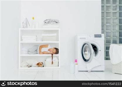 Cozy washing room with washer, sleeping girl with dog on shelf, bottles of liquid powder on floor, basket full of dirty laundry. Child has rest after helping mum to wash clothes. Domestic atmosphere
