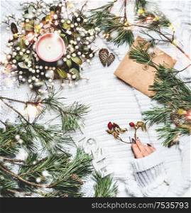 Cozy winter concept. Female hand in warm knitted sweater on white plaid background with pine branches, candles, and cones. Beautiful bokeh of snow. Kraft paper envelope. Frame. Top view. Insta style