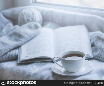 Cozy winter still life: mug of hot tea and book with warm plaid on windowsill against snow landscape from outside.