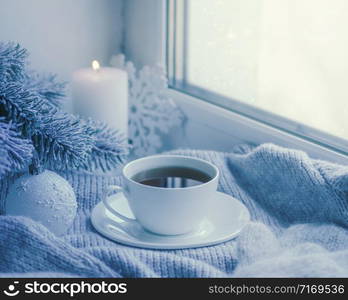 Cozy winter still life: mug of hot tea and book with warm plaid on windowsill against snow landscape from outside.