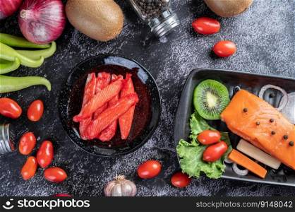 Crab stick put on a black plate with raw salmon, tomatoes, kiwi, shallots and sweet peppers on the black cement floor.