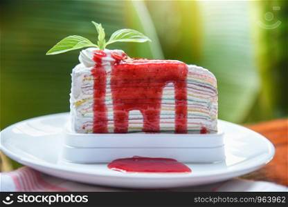 Crape cake slice with strawberry sauce on white plate and nature green background / Piece of cake rainbows with whipped cream