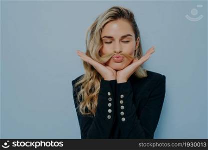 Crazy blonde woman in black blazer having fun and fooling around with closed eyes, using her hair as moustache, making weird faces, supporting head with hands, standing isolated on blue background. Crazy blonde woman having fun and fooling around