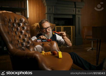 Crazy funny senior businessman playing toys enjoying fun time after hard working day in home office. Elderly male sitting on floor nearby armchair. Mental health and stress relief concept. Funny senior businessman playing toys enjoying fun time after hard working day
