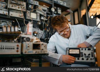 Crazy scientist holds electrical device in laboratory. Electrical testing tools on background. Lab equipment, engineering workshop
