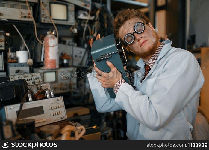 Crazy scientist in glasses holds electrical device in laboratory. Electrical testing tools on background. Lab equipment, engineering workshop