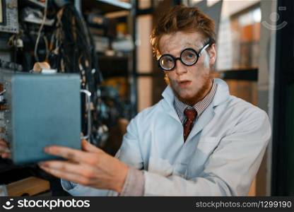 Crazy scientist in glasses holds electrical device in laboratory. Electrical testing tools on background. Lab equipment, engineering workshop. Scientist in glasses holds electrical device