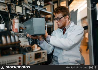 Crazy scientist in glasses holds electrical device in laboratory. Electrical testing tools on background. Lab equipment. Scientist in glasses holds electrical device