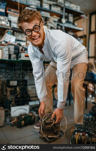Crazy scientist lifts heavy electrical device in laboratory. Electrical testing tools on background. Lab equipment, engineering workshop
