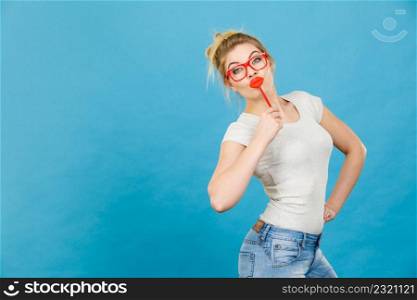 Crazy woman casual style nerdy glasses holding red fake lips on stick having fun, on blue. Photo take and carnival funny accessories concept.. Happy woman holding fake lips on stick