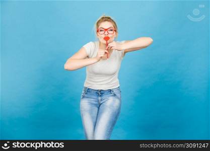 Crazy woman casual style nerdy glasses holding red fake lips on stick having fun, on blue. Photo take and carnival funny accessories concept.. Happy woman holding fake lips on stick