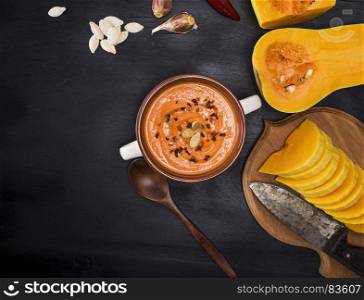 cream fresh pumpkin soup in a round ceramic dish and ingredients for cooking on a black background, empty space on the left