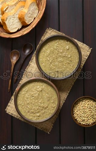 Cream of lentil soup in rustic bowls with wooden spoons, bread slices in basket and raw lentils in small bowl on the side, photographed overhead on dark wooden with natural light