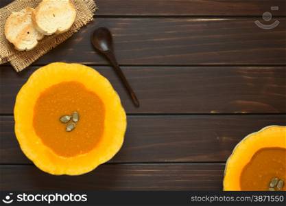 Cream of pumpkin soup served in half pumpkins with pepita seeds on top, toasted bread slices on the side, photographed overhead on dark wood with natural light