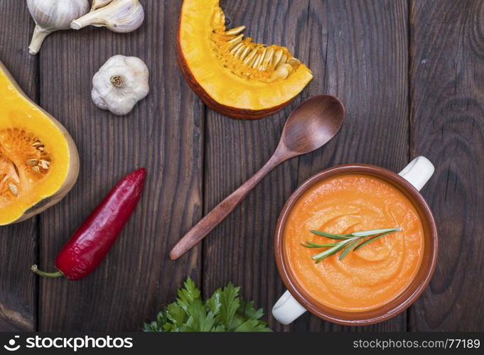 creamy pumpkin soup in a ceramic plate with handles and pieces of fresh pumpkin on a brown wooden table, top view