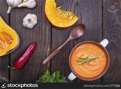 creamy pumpkin soup in a ceramic plate with handles and pieces of fresh pumpkin on a brown wooden table, top view